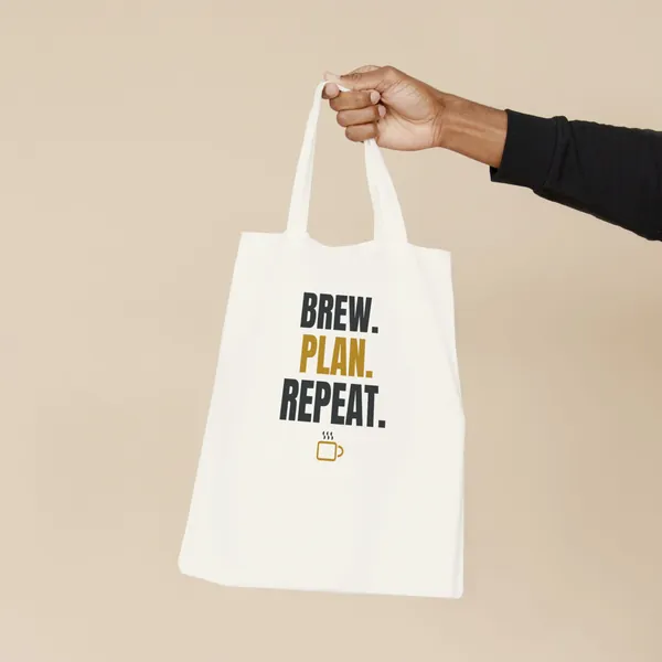 Natural canvas tote bag printed with "Brew. Plan. Repeat.", held by the handles