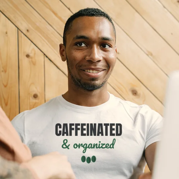 Man wearing a white "Caffeinated and Organized" tee in an office with a wooden wall