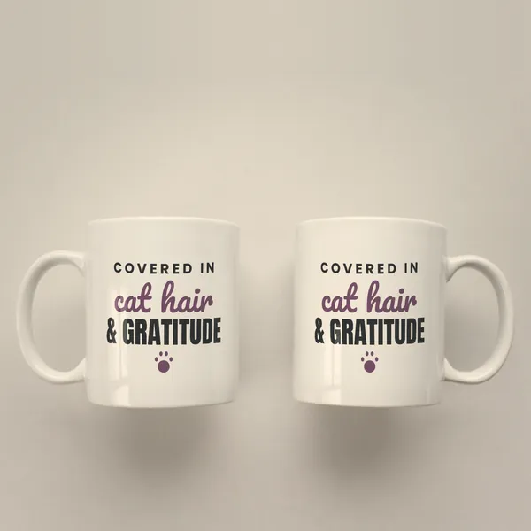 Two white 11 oz ceramic mugs printed with "Covered in Cat Hair and Gratitude", handles left and right