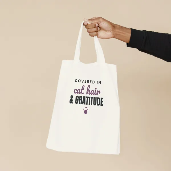 Natural canvas tote bag printed with "Covered in Cat Hair and Gratitude", held by the handles