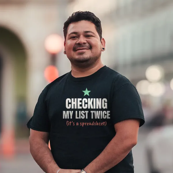 Man wearing a black "Checking My List Twice (It's a Spreadsheet)" tee outdoors on a city street (light-ink print for dark shirts)