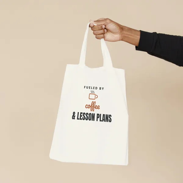 Natural canvas tote bag printed with "Fueled by Coffee and Lesson Plans", held by the handles
