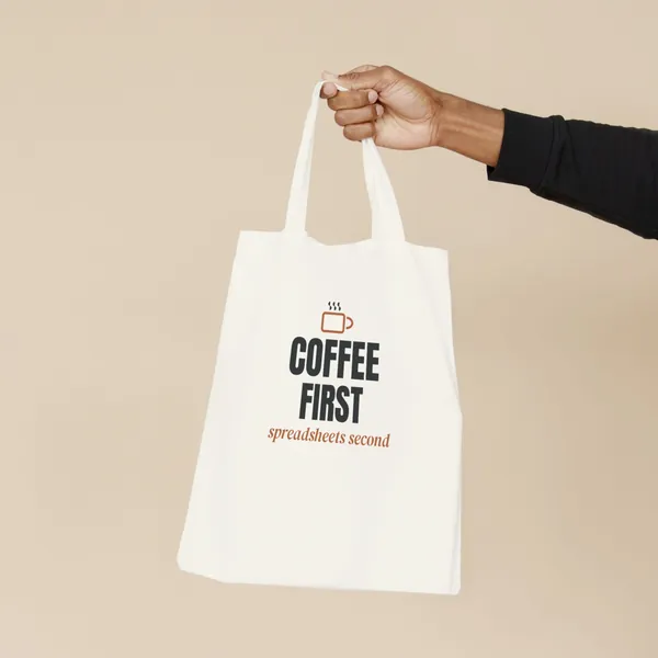 Natural canvas tote bag printed with "Coffee First, Spreadsheets Second", held by the handles