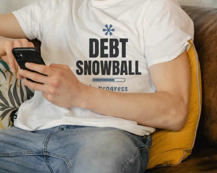Man wearing a white "Debt Snowball in Progress" tee on a couch with his phone