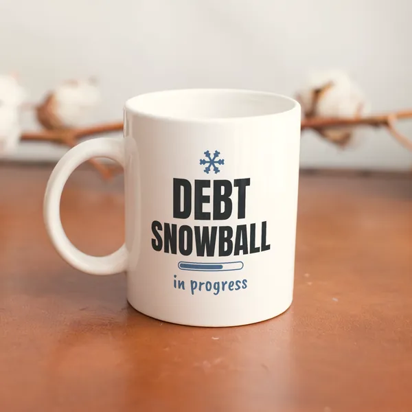White 11 oz ceramic mug printed with "Debt Snowball in Progress" on a wooden table