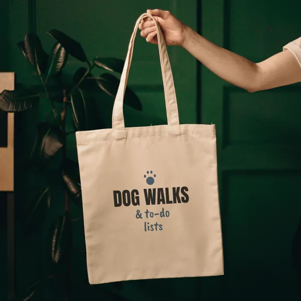 Natural canvas tote bag printed with "Dog Walks and To-Do Lists", held up against a green door
