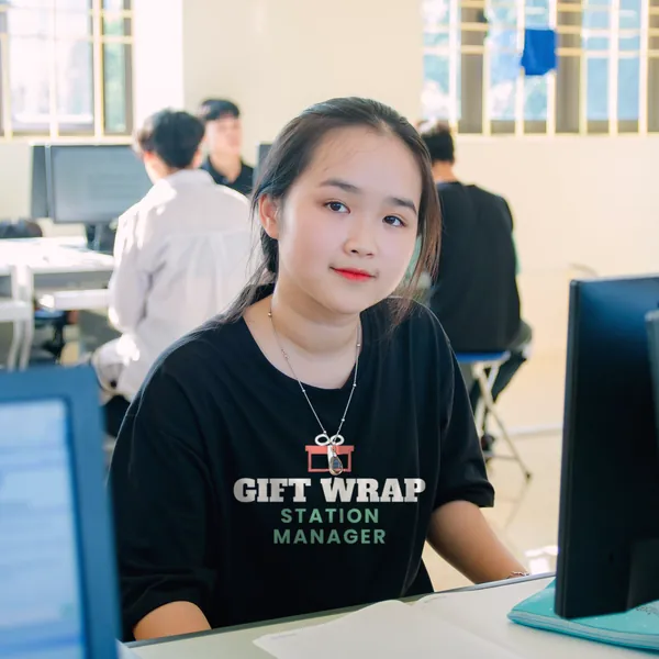 Woman wearing a black "Gift Wrap Station Manager" tee at a desk in a computer lab (light-ink print for dark shirts)
