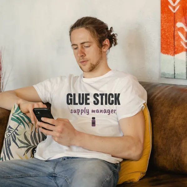 Man wearing a white "Glue Stick Supply Manager" tee on a couch with his phone