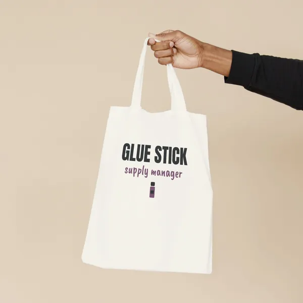 Natural canvas tote bag printed with "Glue Stick Supply Manager", held by the handles