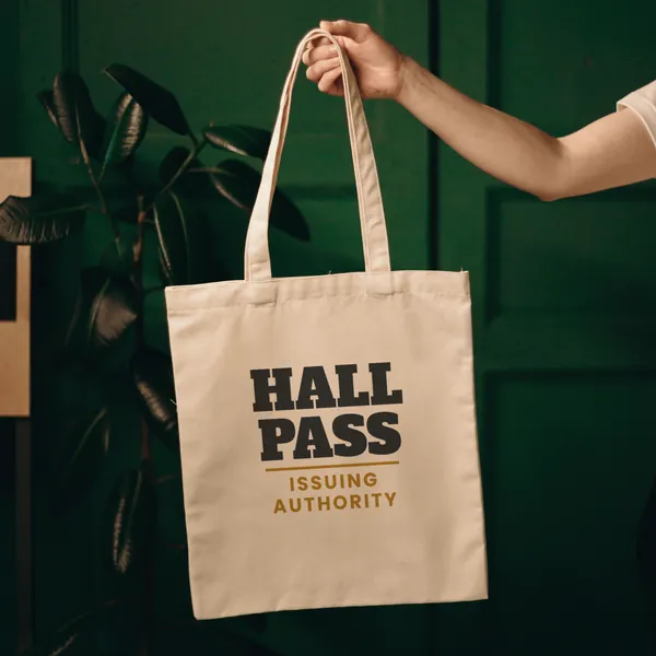 Natural canvas tote bag printed with "Hall Pass Issuing Authority", held up against a green door