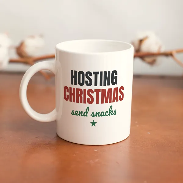 White 11 oz ceramic mug printed with "Hosting Christmas, Send Snacks" on a wooden table