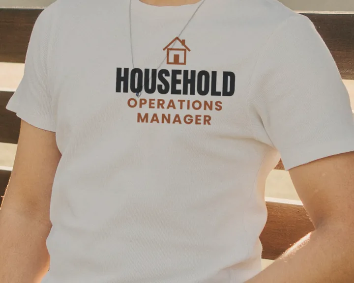 Man wearing a white "Household Operations Manager" tee outdoors by a slatted fence