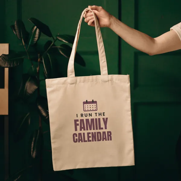 Natural canvas tote bag printed with "I Run the Family Calendar", held up against a green door