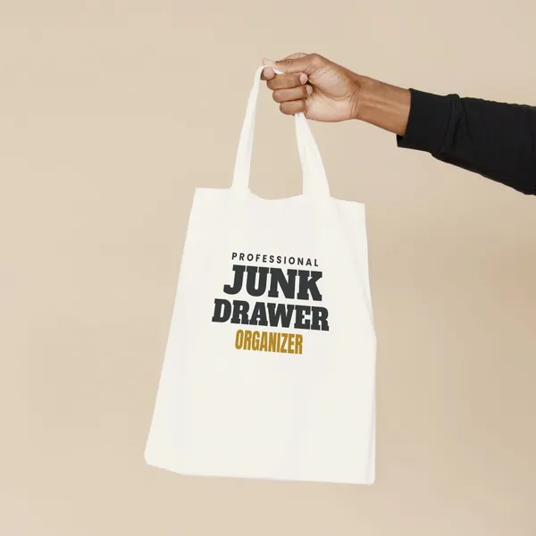 Natural canvas tote bag printed with "Professional Junk Drawer Organizer", held by the handles