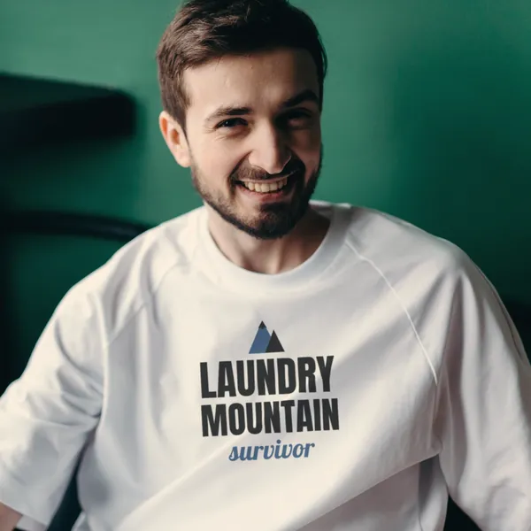 Man wearing a white "Laundry Mountain Survivor" tee at home by a green wall