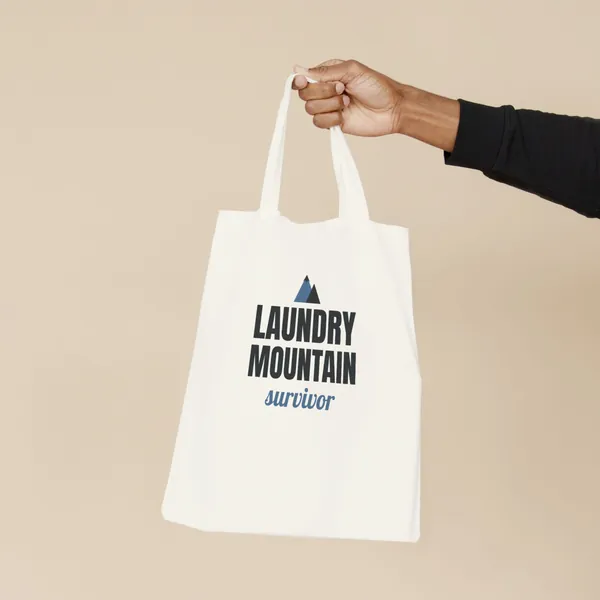 Natural canvas tote bag printed with "Laundry Mountain Survivor", held by the handles