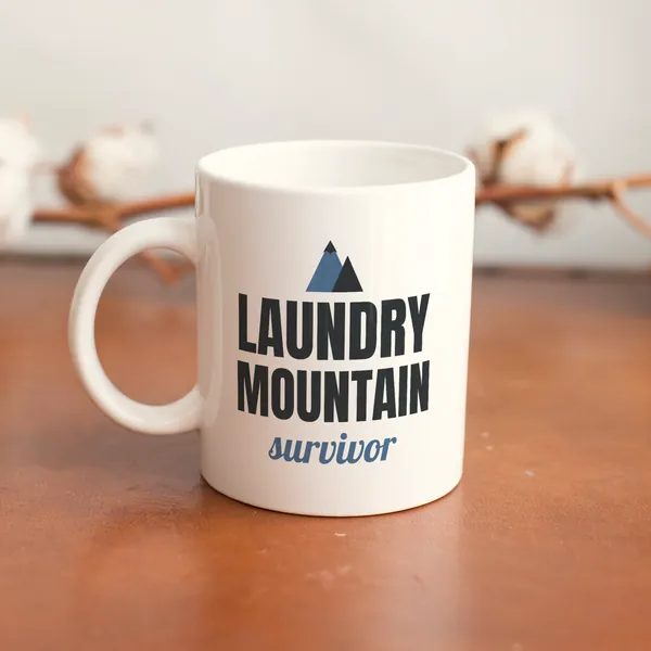 White 11 oz ceramic mug printed with "Laundry Mountain Survivor" on a wooden table