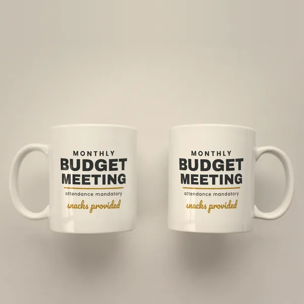 Two white 11 oz ceramic mugs printed with "Monthly Budget Meeting - Snacks Provided", handles left and right