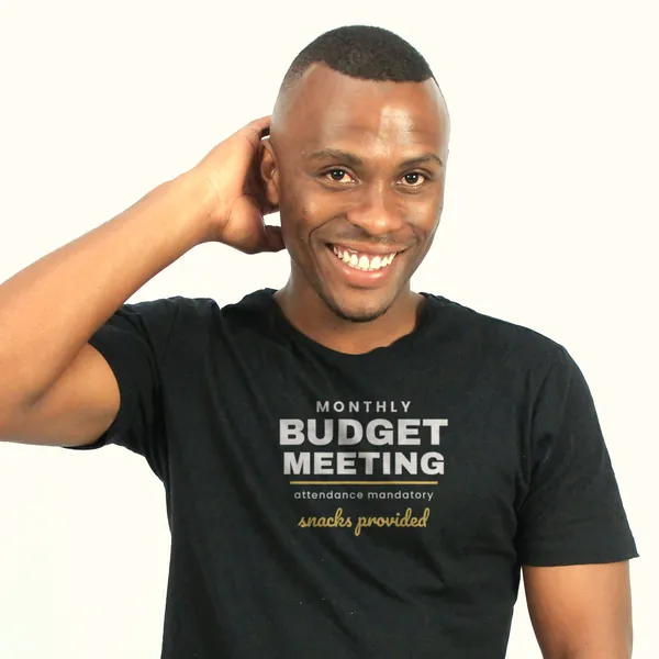 Man wearing a black "Monthly Budget Meeting - Snacks Provided" tee in a bright studio (light-ink print for dark shirts)