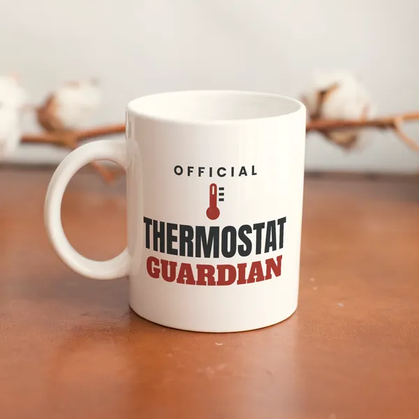 White 11 oz ceramic mug printed with "Official Thermostat Guardian" on a wooden table
