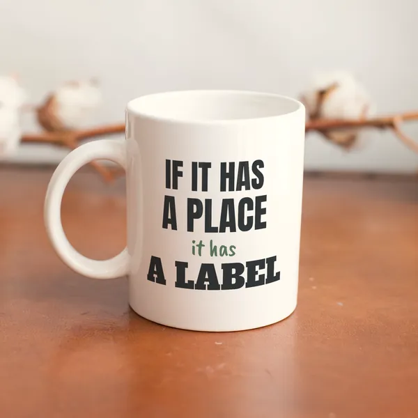White 11 oz ceramic mug printed with "If It Has a Place, It Has a Label" on a wooden table