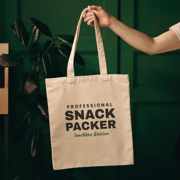 Natural canvas tote bag printed with "Professional Snack Packer", held up against a green door