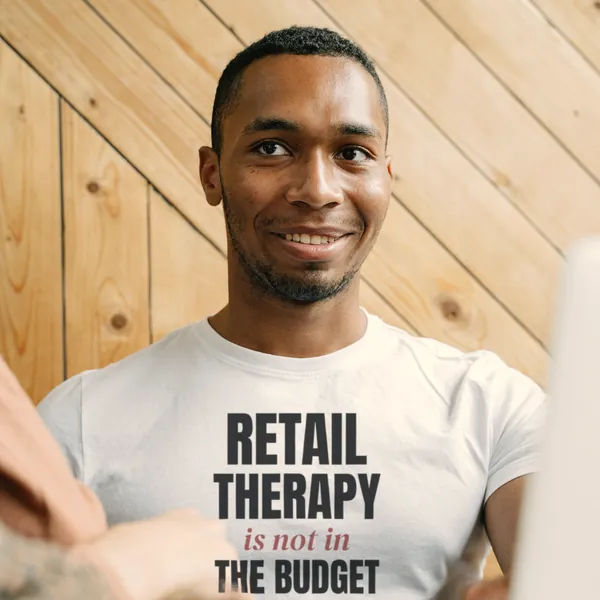 Man wearing a white "Retail Therapy Is Not in the Budget" tee in an office with a wooden wall