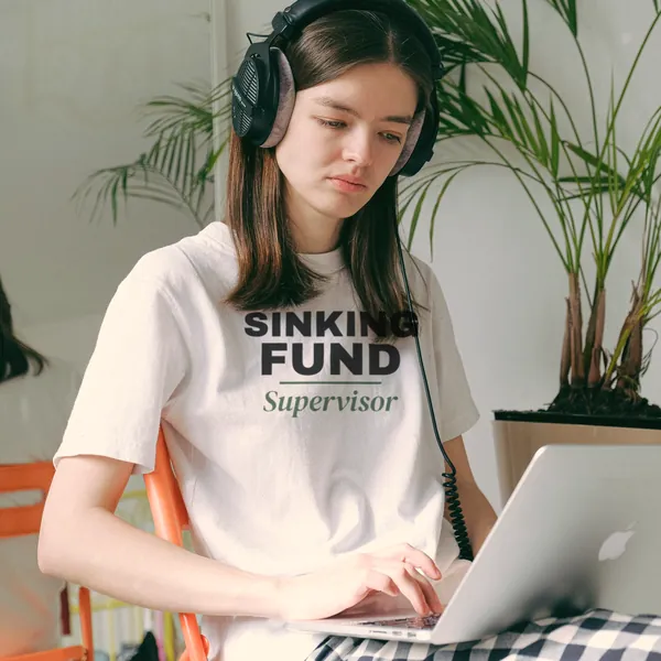 Woman wearing a white "Sinking Fund Supervisor" tee working on a laptop in a home office