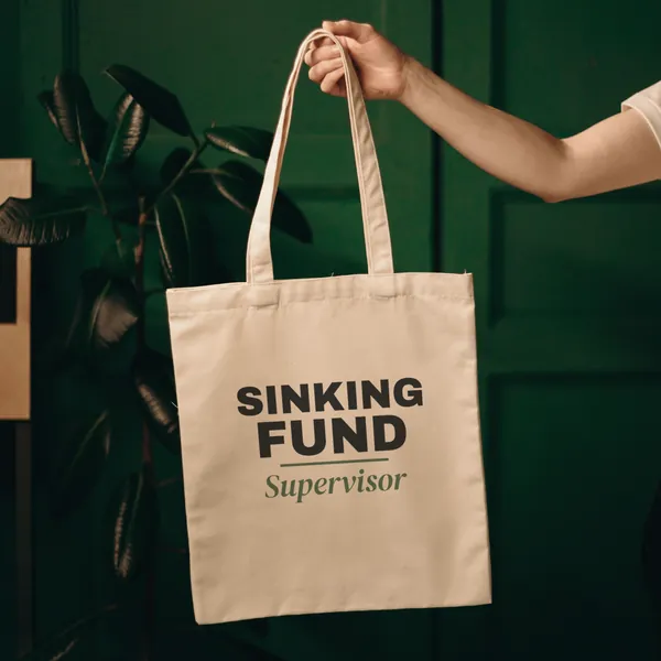 Natural canvas tote bag printed with "Sinking Fund Supervisor", held up against a green door